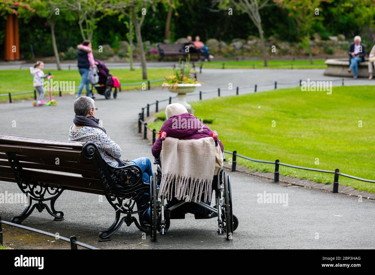 Ältere Person im Rollstuhl mit Wächter auf einer Bank im St. Stephen`s Green Park in Dublin während der Covid-19 Pandemic Ireland. Stockfoto